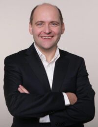 Speaker Dr. Gregor Wittke Ein Mann in einem schwarzen Anzug und weißem Hemd, mit verschränkten Armen und einem freundlichen Lächeln, vor einem neutralen Hintergrund.