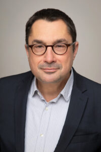 Speaker Dr. Liviu-Mihai Blaga; Selbstporträt eines Mannes mit Brille in einem eleganten Anzug, vor einer schlichten, weißen Wand.