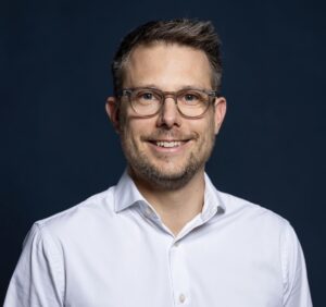 Speaker Benjamin Waschow. Ein Mann mit Brille und freundlichem Lächeln, im weißen Hemd vor einem dunkelblauen Hintergrund. Der Kontrast hebt die Person hervor und gibt dem Bild Tiefe.