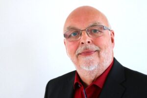Portaitfoto: Ein glatzköpfiger Mann mit leichtem Bart und Brille, in rotem Hemd gekleidet, schaut freundlich in die Kamera.