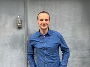 Ein Mann in einem blauen Hemd steht vor einer grauen Wand. Das Foto zeigt ihn bis zur Hüfte. Er schaut freundlich in die Kamera.
